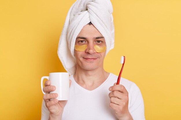위생적인 아침 일과와 치아 미백 침착한 잘생긴 남자는 양치질을 하고 흰색 컵을 손에 들고 머리에 수건을 씁니다.
