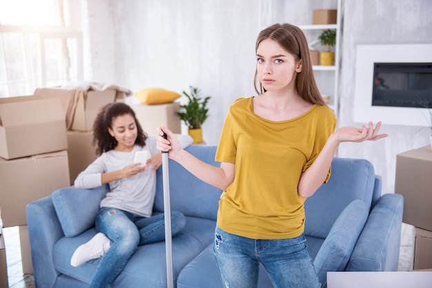 I am angry. Beautiful young girl cleaning the floor and being mad at her lazy roommate while she sitting texting on the sofa instead of helping her