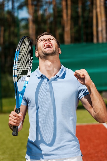 Sono il migliore! felice giovane uomo in polo tenendo la racchetta da tennis e gesticolando mentre si sta in piedi sul campo da tennis