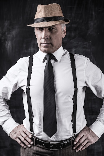 Photo i am a boss! confident senior man in hat and suspenders looking at camera and holding hands on hip while standing against dark background