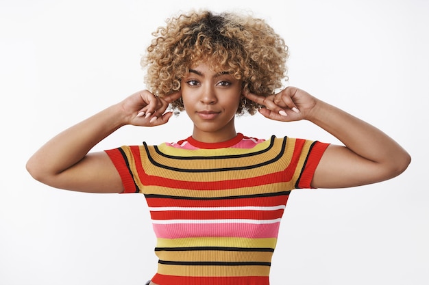 I am not listening to your bullshit. Stylish self-assured good-looking african american female with fair haircut close ears with index fingers and smiling as unwilling listen over white wall