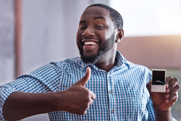 I am overjoyed. Happy young African man holding an engagement ring preparing for proposal and making approval gesture.