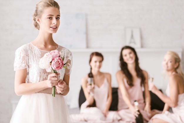 I am so excited. Concentrated shy young bride standing in the white colored bedroom while holding the wedding bouquet and her bridesmaids having fun