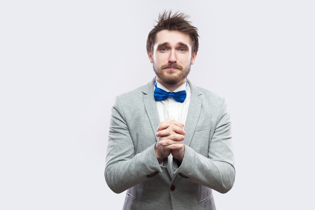 I am sorry, please forgive me. sad handsome bearded man in casual grey suit and blue bow tie looking at camera and asking for help or forgivness. indoor studio shot, isolated on light grey background.