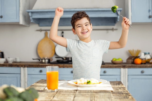 私は強いです。健康的な朝食を食べて、笑顔で彼の筋肉を見せているハンサムな陽気な黒髪の少年