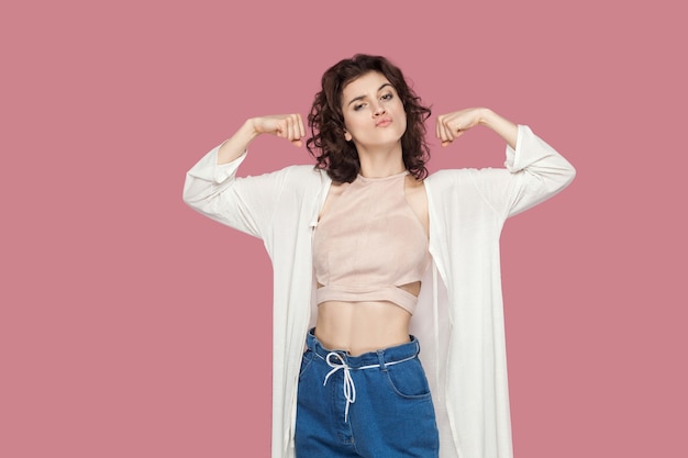 I am strong and independent. Satisfied beautiful brunette young woman with curly hair in casual style standing with strong gesture and looking at camera. indoor studio shot isolated on pink background