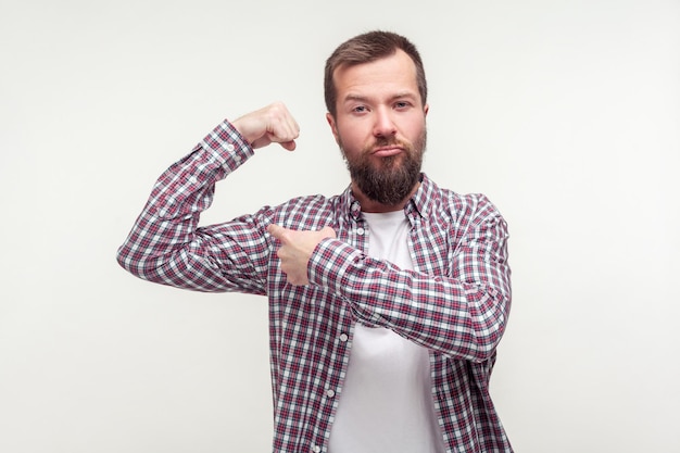 I am strong Portrait of proud bearded man in casual plaid shirt pointing at raised hand demonstrating biceps confident in body feeling powerful indoor studio shot isolated on white background