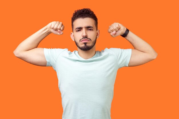 I am strong and powerful Portrait of confident successful brunette man with beard in white tshirt showing biceps full of motivation and willpower indoor studio shot isolated on orange background