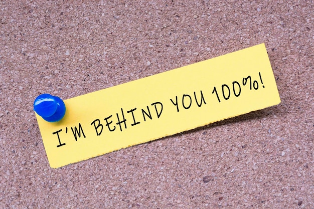 I am behind you 100 percent words on stick note and blue pinned to a cork notice board