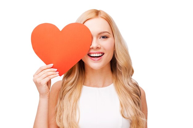 I believe in love! Beautiful young blond hair woman holding heart shaped valentine card in front of her eye and smiling while standing isolated on white