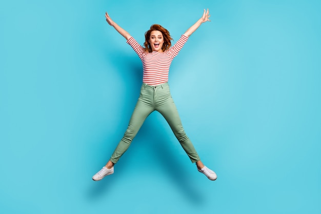 I ben beschikbaar! Full body foto van gekke dame springen hoog verheugend weekend zonnige dag gespreide handen dragen casual rood wit overhemd groene broek schoenen geïsoleerde blauwe kleur