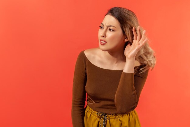 I can\'t hear. portrait of pretty woman in brown blouse holding\
hand near ear, listening attentively with interest private\
conversation, confidential talk. indoor studio shot isolated on red\
background