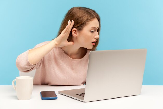 I can't hear, Woman holding arm near ear trying to listen secret talk on video call on laptop, bad internet connection, online conference from home office. indoor studio shot isolated, blue background