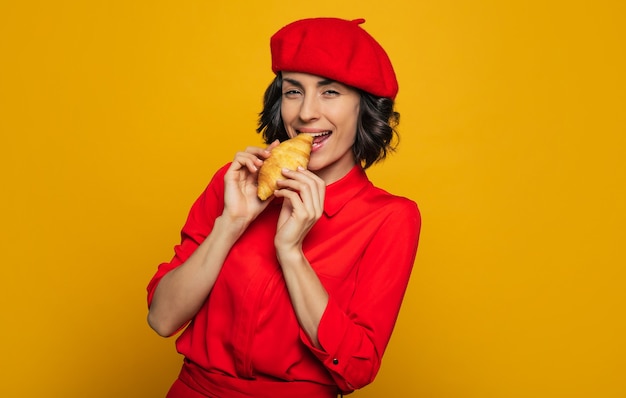 Фото Я не могу устоять перед кусочком французского печенья! половинная фотография молодой француженки, одетой в парижском стиле, с красным беретом, откусывающая круассан.