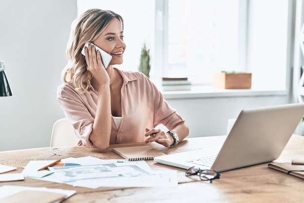 I can totally make it work! Beautiful young woman in smart casual wear smiling and talking on the phone while sitting in modern office