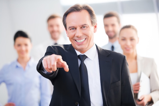 I choose you! Confident mature businessman pointing you and smiling while his colleagues standing in the background