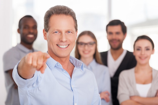 I choose you! Confident mature man pointing you and smiling while group of people in casual wear standing on background