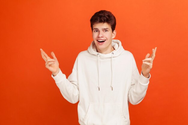 I don39t know Extremely surprised young man in casual style hoodie spreading his hands Shocked with amazing news Indoor studio shot isolated on orange background