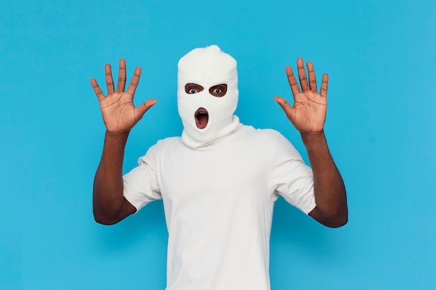 I give up shocked african american male thug in white balaclava raising hands on blue background