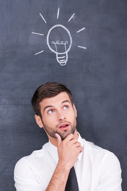 I got it. Thoughtful young man holding hand on chin and looking up while standing against blackboard with chalk drawing of light bulb