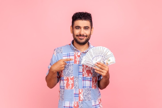 У меня большие деньги. Портрет богатого молодого взрослого человека, указывающего на долларовые банкноты и смотрящего со счастливым выражением, финансовые сбережения. Крытая студия снята на розовом фоне.
