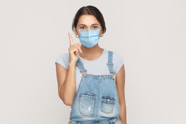 Ho un'idea. ritratto di giovane donna bruna felice con maschera medica chirurgica in tuta di jeans in piedi sorpresa e guardando la telecamera sorridendo. studio al coperto colpo isolato su sfondo grigio.