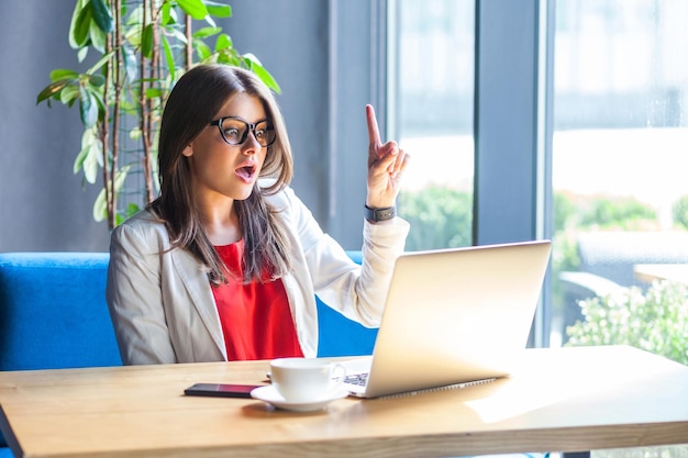 I have an idea portrait of surprised beautiful stylish brunette\
young woman in glasses sitting looking at her laptop screen with\
idea gesture and amazed indoor studio shot cafe office\
background