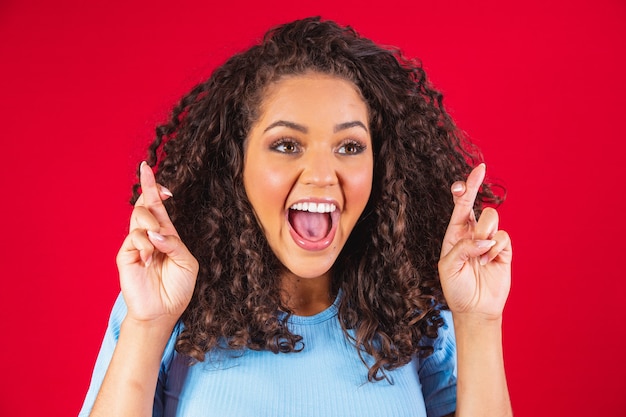 I have to win. Joyful young brunette female clenches teeth, raises fingers crossed, makes desirable wish, curly hair, mixed race, African, Brazilian girl, waits for good news, stands over red wall