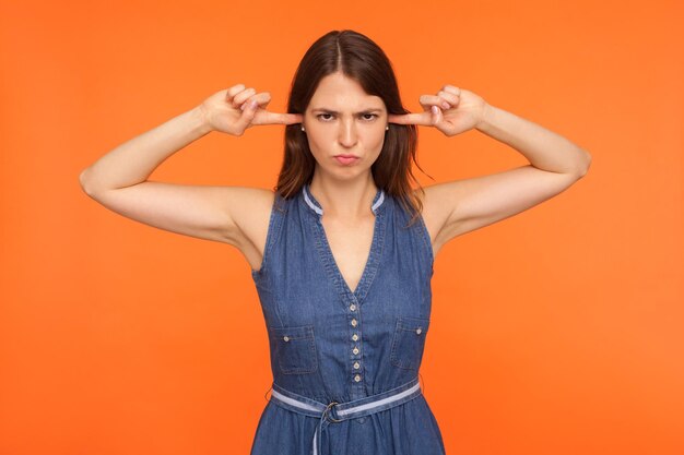 I don't hear. Annoyed brunette woman in denim dress closing ears with fingers and looking angrily to camera, ignoring talk, not listening to other opinions. studio shot isolated on orange background