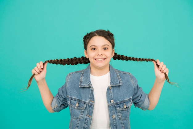 I just let my hair go. Happy girl hold long hair blue background. Small child wear braided hair style. Little hair model with beauty look. Hairdressing salon. Kids barber. Haircare products.