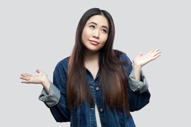 I don't know. Confused beautiful brunette asian young woman in casual blue denim jacket with makeup standing raised arms and looking away thoughtful. studio shot, isolated on light grey background.