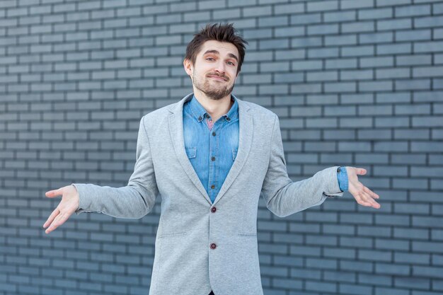 I don't know. Portrait of confused handsome bearded man in casual style standing, raised arms and looking at camera with doubtful face. indoor studio shot on brick wall background.