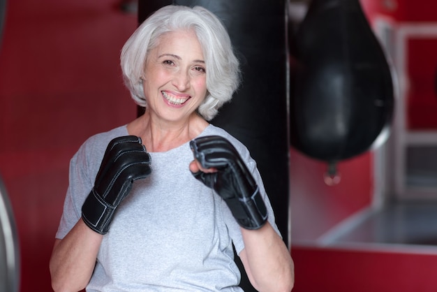 I like box. Delighted pretty senior woman smiling and holding hands like boxer while training in a gym.an holding hands like a boxer.