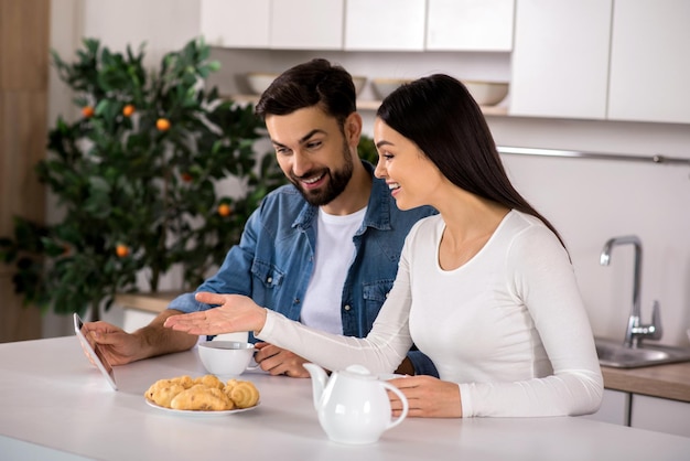 Photo i like it cheerful nice couple using tablet while sitting at the table in the kitchen