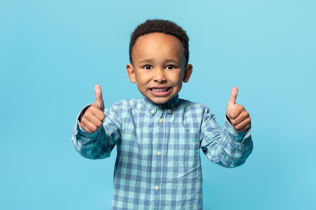 I like it Happy african american adorable boy showing thumbs up and widely smiling posing over blue background