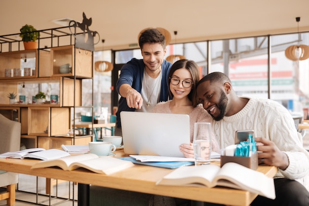 I like it. Young handsome man is pointing at laptop screen while while sitting at cafe and studying with his two fellows.