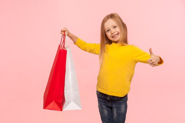 I like shopping at kids store. Portrait of happy carefree little girl holding large packages and showing thumb up, satisfied with Black Friday discounts. indoor studio shot isolated on pink background