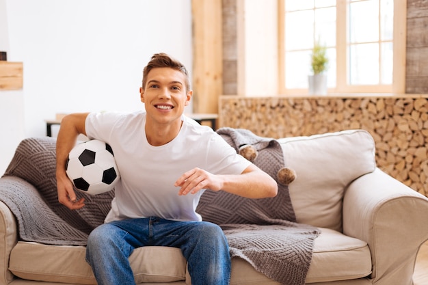 I love football. Good-looking cheerful well-built adolescent smiling and holding a soccer ball while sitting on the couch