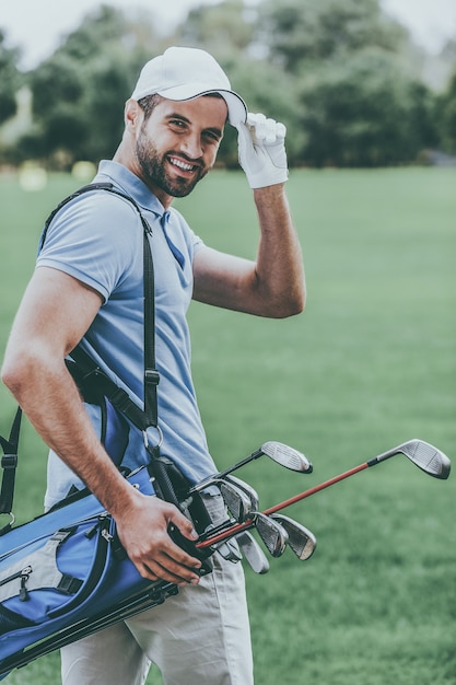 Я люблю играть в гольф! Вид сзади молодого счастливого гольфиста, несущего сумку для гольфа с водителями и смотрящего через плечо, стоя на поле для гольфа