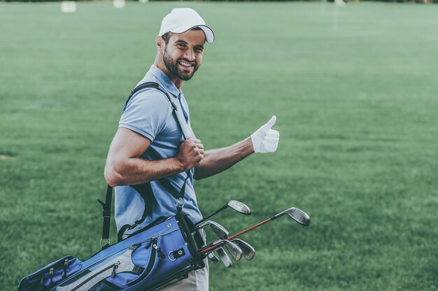 I love golfing! rear view of young happy golfer carrying golf\
bag with drivers and looking over shoulder while standing on golf\
course