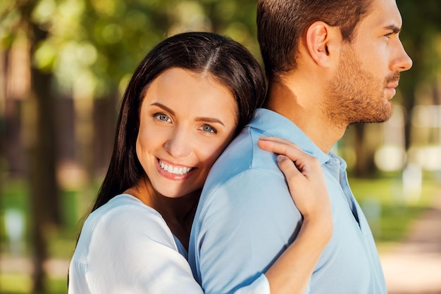 I love him so much! Beautiful young woman leaning to the back of her boyfriend and smiling at camera while both standing outdoors