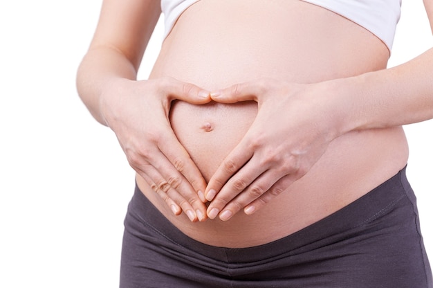 I love my baby. Cropped image of pregnant woman holding hands in heart shape on her abdomen while standing isolated on white