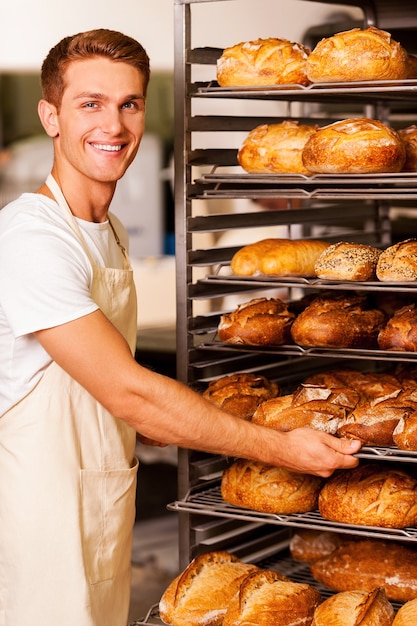 I love my job! Handsome young baker in apron taking fresh baked bread from the tray and smiling