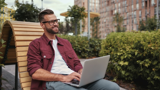 Amo il mio lavoro felice giovane bell'uomo con la barba in abiti casual e occhiali da vista che lavora al computer portatile e sorride mentre è seduto sulla panchina all'aperto. bloggare. vita di città