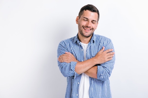 I love myself! Portrait of egoistic brunette man with beard standing with closed eyes, embracing himself and smiling form pleasure and proud. Indoor, isolated on white background