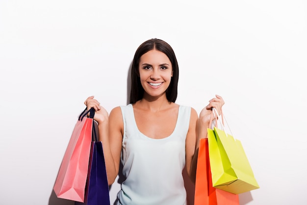 I love shopping! Attractive young woman in dress carrying colorful shopping bags and smiling while standing against white background