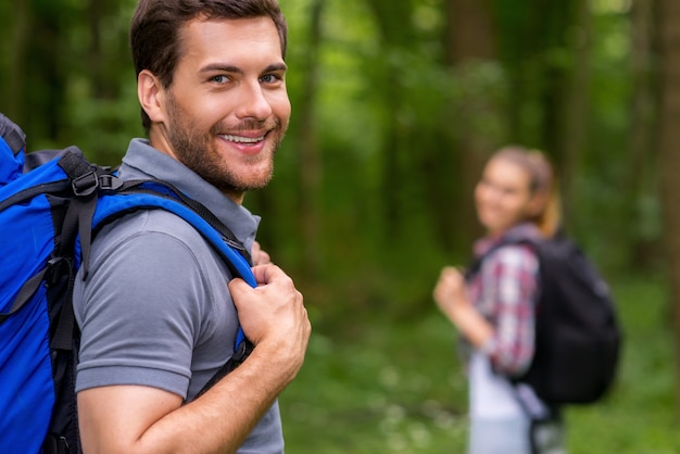 私は旅行が大好き。バックパックのハンサムな若い男が肩越しに見て、バックグラウンドで女性と森を歩きながら笑っている