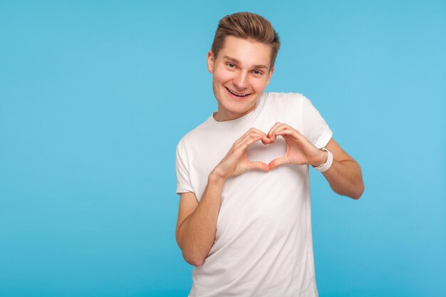 I love you. Portrait of happy man in white t-shirt expressing feelings with amorous heart gesture near chest, romantic symbol of affection, devotion. indoor studio shot isolated on blue background