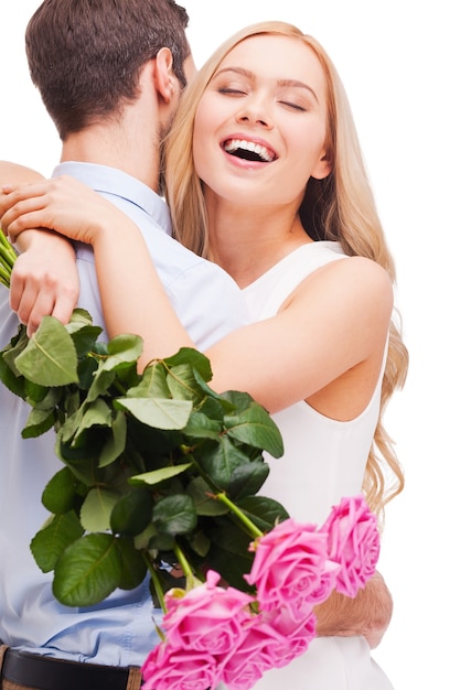 Я так сильно тебя люблю! Красивая молодая влюбленная пара обнимается, пока женщина держит букет розовых роз и улыбается, и оба стоят, изолированные на белом фоне