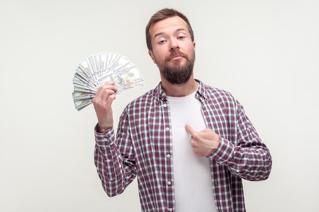 I made money myself. Portrait of arrogant bearded man in casual plaid shirt pointing at himself and holding dollars, proud of big earnings, startup. indoor studio shot isolated on white background